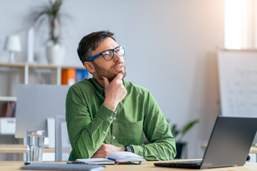 Focused Caucasian Man In Green Shirt Thinking While Working On Laptop At Modern Office, Sitting At Desk With Notebook And Glass Of Water. Inspiring Business Concept © Prostock-studio