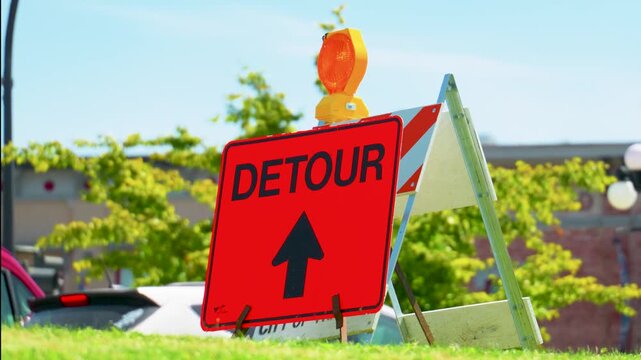 Bright orange diamond detour sign with black up arrow mounted on a wooden barricade with an orange safety flasher