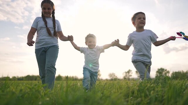 Toddler walks forward in bright sunlight holding girl hand, while boy steps beside with toy. Outdoor field stretches around them. Walk continues with toddler supported by hand of girl and boy.
