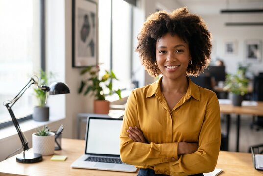  Casual portrait of a successful young African American female creative director standing by her desk in a contemporary startup office.