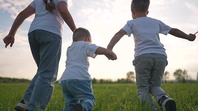 Children walk together on grassy field. Girl holds toddler hand. Boy walks with windmill toy. Toddler steps forward in outdoor nature. Sibling walk in field. Walk in nature with toddler outdoors
