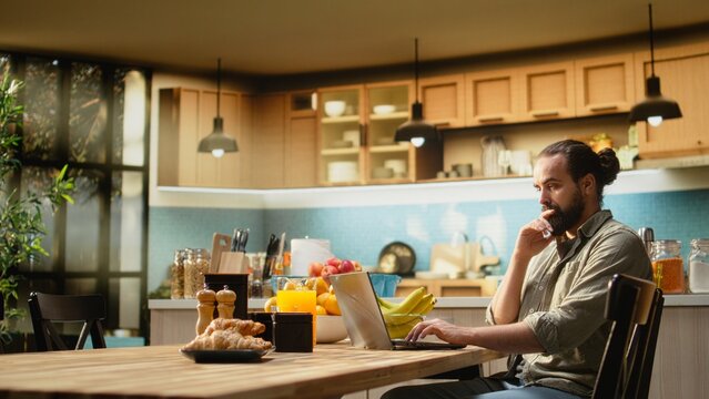 Uncertain freelancer being confused about ambiguous tasks at home, remote work on laptop in the kitchen, Disoriented disorganized man unsure about telecommuting job, frowning.
