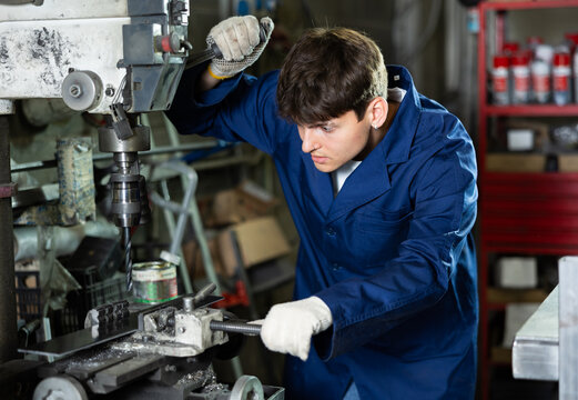 Skilled young factory worker, in blue work coat and protective gloves, working on drilling machine in processing workshop, performing holes in metal part