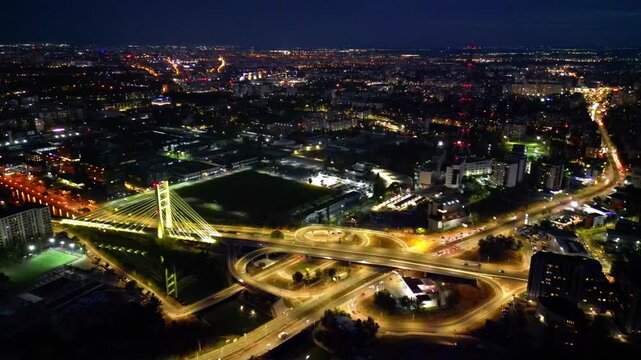 Cinematic high angle night view of the illuminated Ciurel bridge in Bucharest, Romania. Modern cable-stayed overpass surrounded by vibrant city traffic lights.