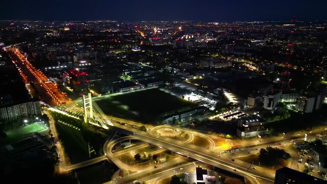 Cinematic high angle night view of the illuminated Ciurel bridge in Bucharest, Romania. Modern cable-stayed overpass surrounded by vibrant city traffic lights.