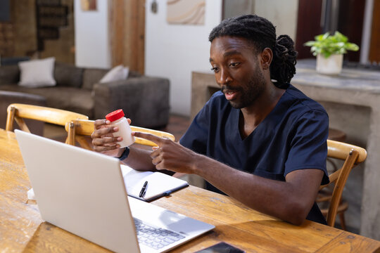 African American man in navy scrubs pointing at pill bottle while referencing open laptop at home