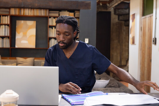 Man in navy medical scrubs sitting at home reviewing laptop, documents, notebook, takeaway cup