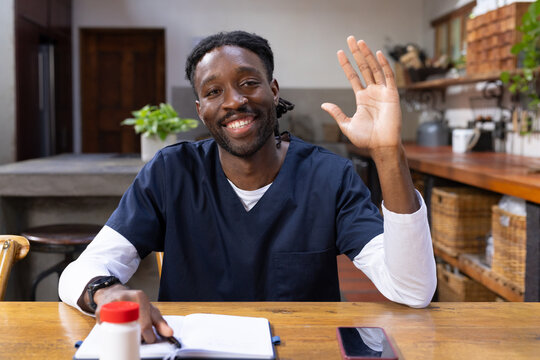 African American man wearing navy scrubs waving hand while sitting at kitchen table with notebook