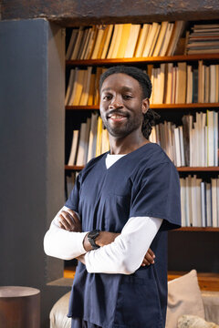 Adult African man standing with arms crossed in study wearing navy scrubs and watch by bookshelf