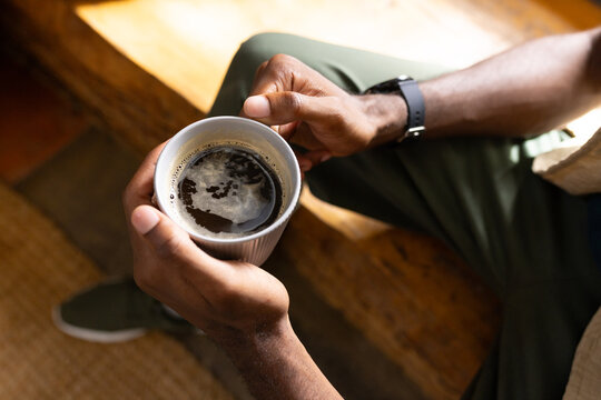 Adult African man cupping ribbed ceramic mug wearing black watch sitting on wooden bench at home