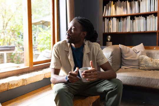 African man sitting at home on bench by wood-framed window holding ceramic mug, looking outside