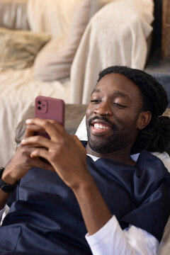 African man reclining on sofa, holding mauve smartphone and smiling, wearing navy top, wristwatch
