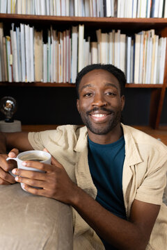 African man wearing beige shirt over teal tee, holding white mug on light sofa, bookshelf behind
