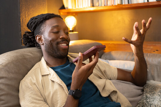 African man reclining on gray couch at home holding smartphone wearing beige shirt and smartwatch