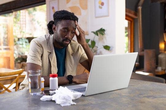 African American man sitting at home dining table holding head using laptop with tissues pills