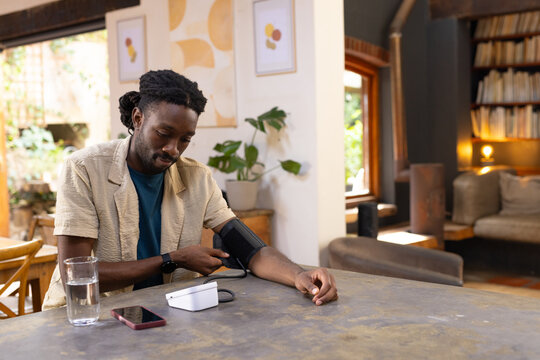 African American man sitting at home wearing blood pressure cuff on left arm using white monitor