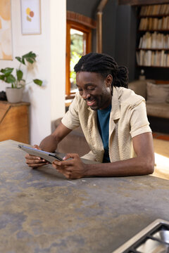 Man with dreadlocks wearing beige shirt over teal tee sitting at home countertop, holding tablet