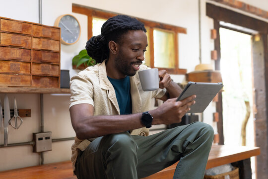 Man sitting at rustic kitchen bench wearing beige shirt holding tablet and sipping mug