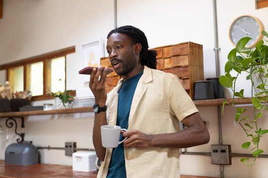 African man standing in home kitchen holding smartphone to mouth and ceramic mug