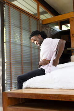 Adult male sitting on platform bed in cabin, lifting shirt and holding lower back, watch visible
