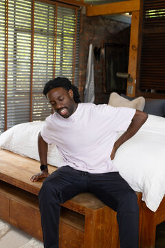 Adult African man sitting on bed edge clutching lower back in bedroom wearing pink T-shirt