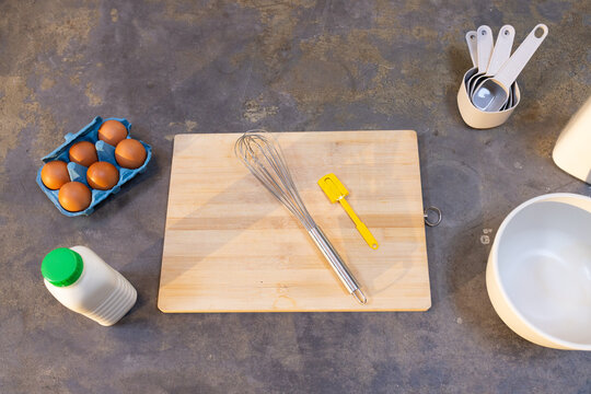 Wooden cutting board is resting with whisk and yellow spatula on dark stone countertop, eggs nearby