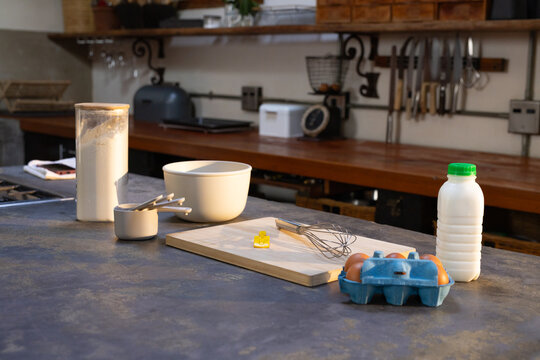 Flour container, measuring cups, mixing bowl, whisk and milk bottle are sitting on stone counter