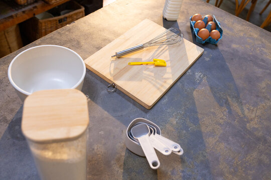 Wooden cutting board is holding whisk and yellow scraper on worn countertop near flour jar, eggs