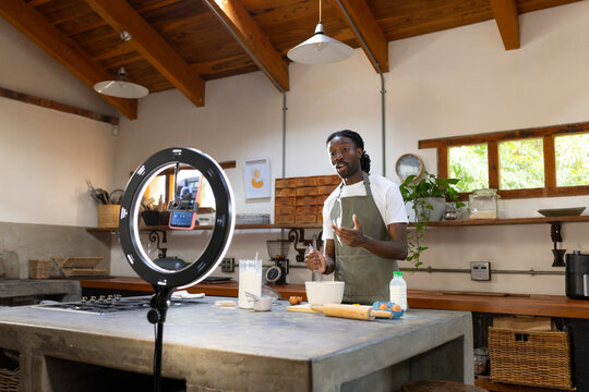 Man wearing green apron mixing batter and talking to smartphone ring light at rustic kitchen island