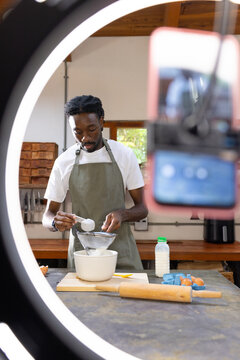 African American man wearing green apron sifting with sieve at kitchen counter filming with phone
