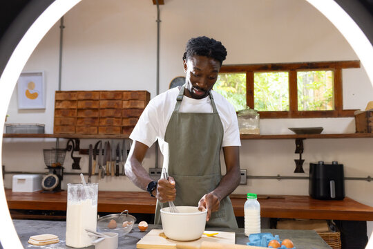 African American man in apron whisking batter with whisk at wooden countertop in bright kitchen