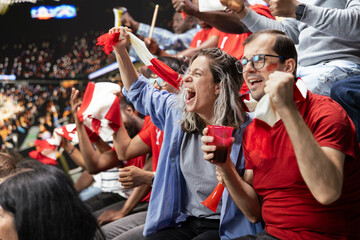 Fototapeta premium Excited couple of football supporters celebrating in stadium stands with red scarf and vuvuzela during an intense live soccer match. Stadium fans celebrate with raised fists, waving scarves.