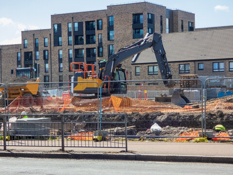 A residential construction site in Scotland, A modern brick apartment building in the background. An excavator and dump truck  is visible, surrounded by temporary fencing and orange safety netting.
