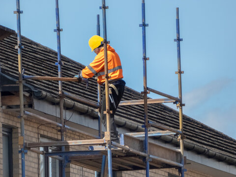 Construction worker is wearing a high-visibility orange jacket and a yellow hard hat.They are performing repairs on a tiled roof while supported by scaffolding. Safe working practices at height. 
