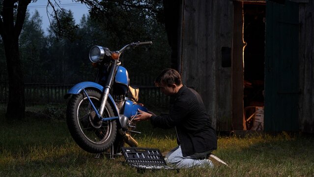 Young man kneeling in a grassy field, adjusting an old blue motorcycle engine during twilight, with a tool kit on the ground and a wooden shed in the background