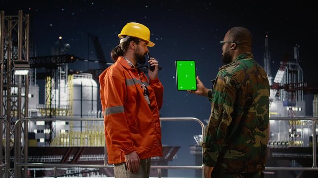 Green screen tablet used by military man on drilling rig with walkie talkie enforcing blockade while a soldier uses mockup device and handheld radio on offshore platform during oil crisis.