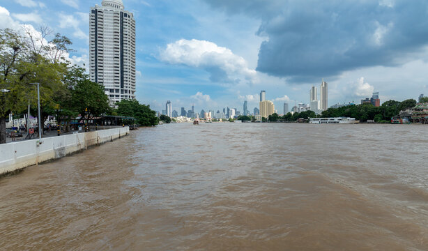 A wide view of the Chao Phraya River in Bangkok, Thailand, with a modern cityscape and skyscrapers in the background under a cloudy sky.