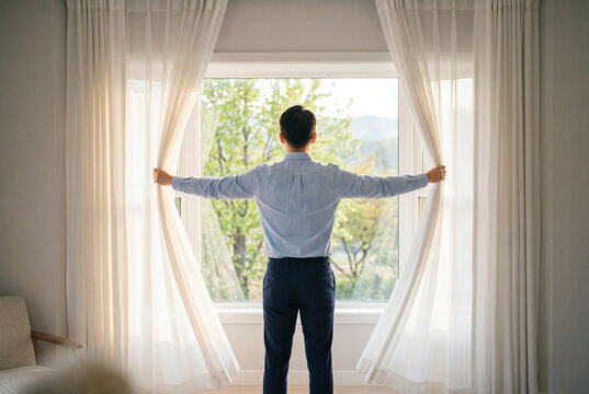 Young man standing backwards and opening window curtains in a bedroom to reveal morning sunlight