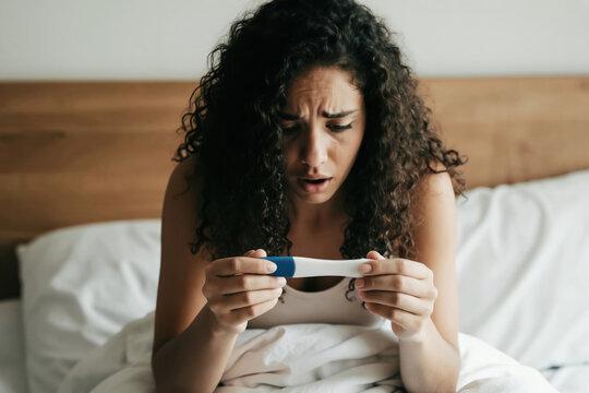 Shocked young woman looks at positive pregnancy test result sitting on bed. Female experiences surprise, worry about becoming mother. Woman checks fertility test in bedroom, emotional reaction to