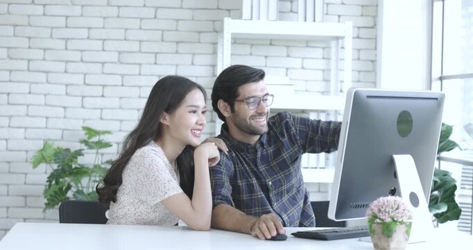 Happy millennial couple in modernn office watching video on computer together, smiling young husband and wife at home browsing Internet using modern computer device.