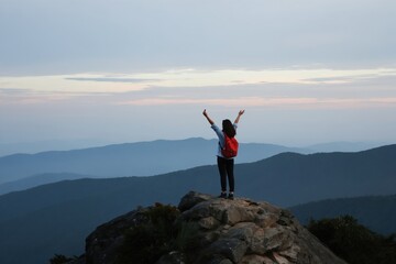 Obraz premium Woman with red backpack stands on mountain peak arms raised in victory. Hiker enjoys sunrise view over misty blue hills. Solo traveler celebrates achievement in nature.
