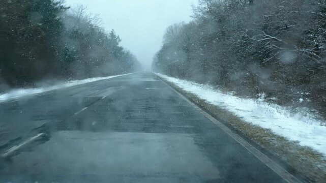 Vehicle driving on empty asphalt road during snowfall. Snow is falling on an empty asphalt road bordered by trees, creating a serene winter landscape as a vehicle navigates the wintry conditions