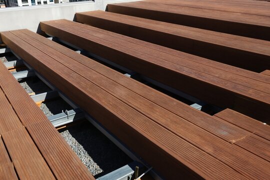 New wooden deck boards laid on metal substructure. Outdoor terrace construction uses brown timber planks. Urban building background shows construction process for new exterior living space.