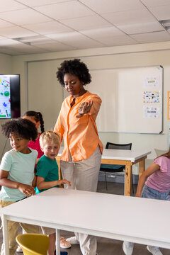 African American teacher in orange top pointing, instructing children at table by whiteboard screen