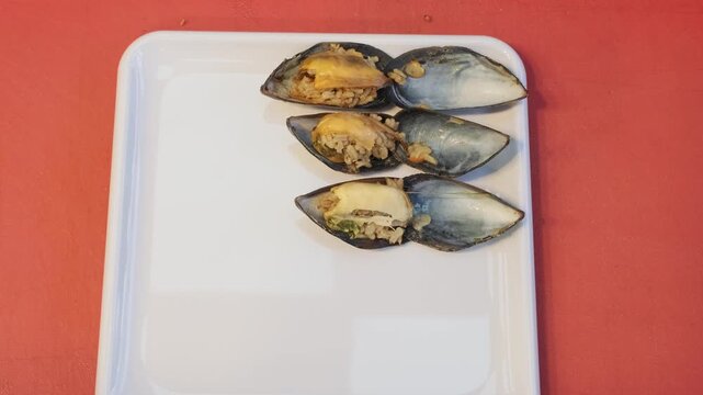 A direct ninety degree overhead close up video of hands neatly arranging open stuffed mussels with seasoned rice on a white square plate, showcasing a popular authentic Turkish seafood snack.