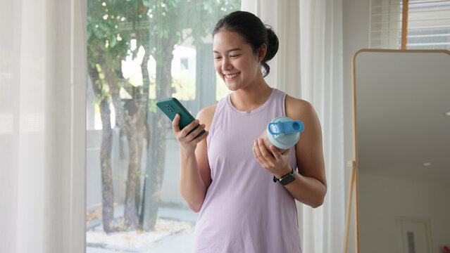 Young gen z active woman drink whey creatine shake bottle eating diet food using phone calorie plan app after home sport workout. health care asia people happy relax protect aging life weight loss.