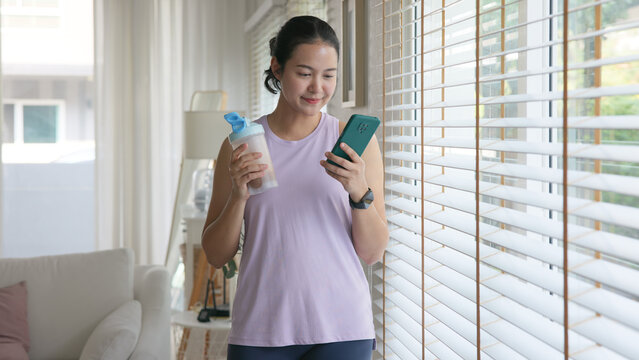 Young gen z active woman drink whey creatine shake bottle eating diet food using phone calorie plan app after home sport workout. health care asia people happy relax protect aging life weight loss.