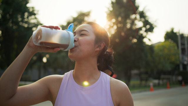 Young active Gen Z woman drink whey boost up protein shake bottle eating diet food calorie plan gain build muscle after workout green park. Health care asia people happy anti aging life weight loss.