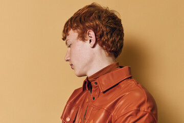 Redhead man in orange leather jacket looking down with neutral expression against beige background. Side profile portrait showing hairstyle and casual fashion details in studio lighting. © SHOTPRIME STUDIO