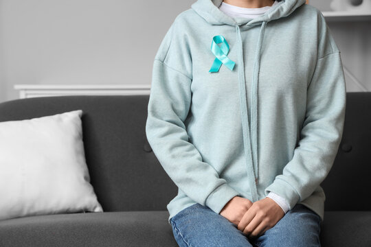 Young woman with turquoise ribbon sitting on sofa at home. Cervical Health Awareness Month